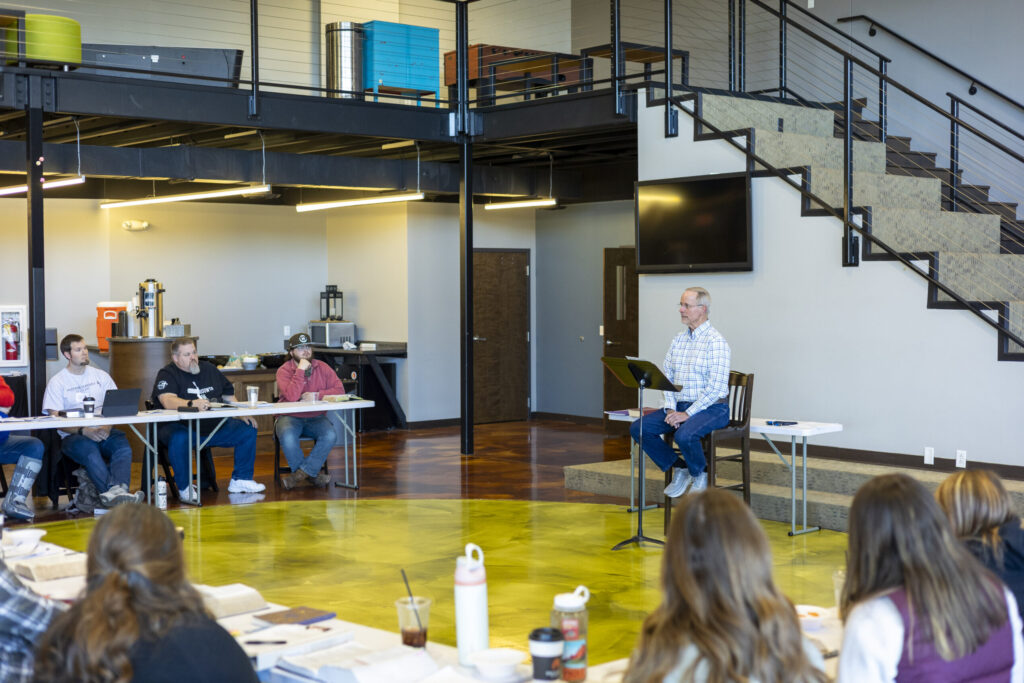 A man speaks to a seated group in a modern meeting space with an upstairs mezzanine and staircase